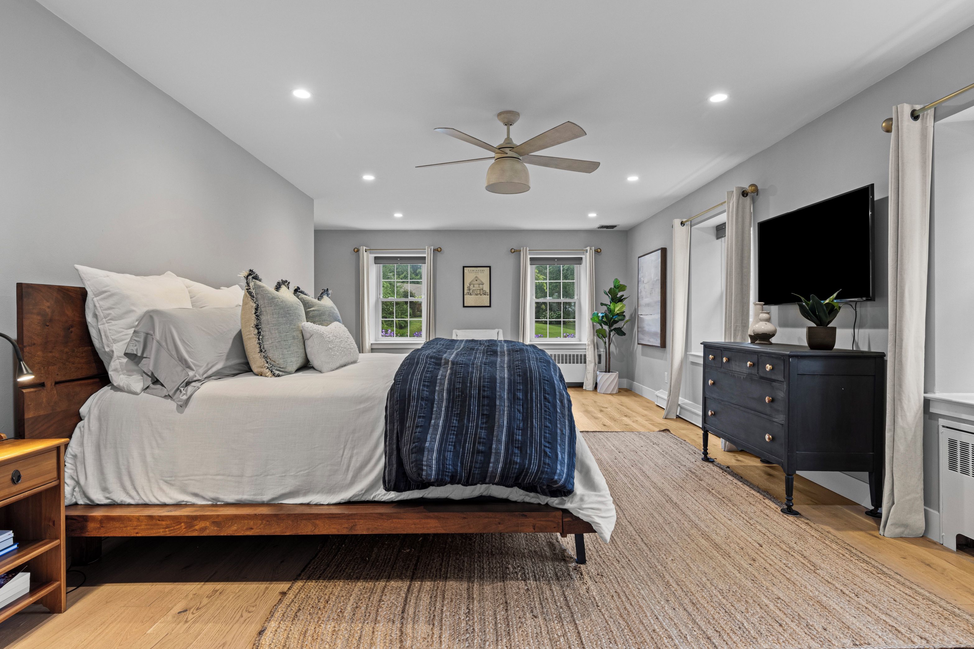 A bedroom with a bed, a ceiling fan, a dresser, a TV, and windows.