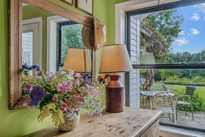 A room with a wooden table, a vase of flowers, a lamp, and a window with a view of greenery. - Woodstock Vermont Escape with Space to Slow Down (Woodstock)