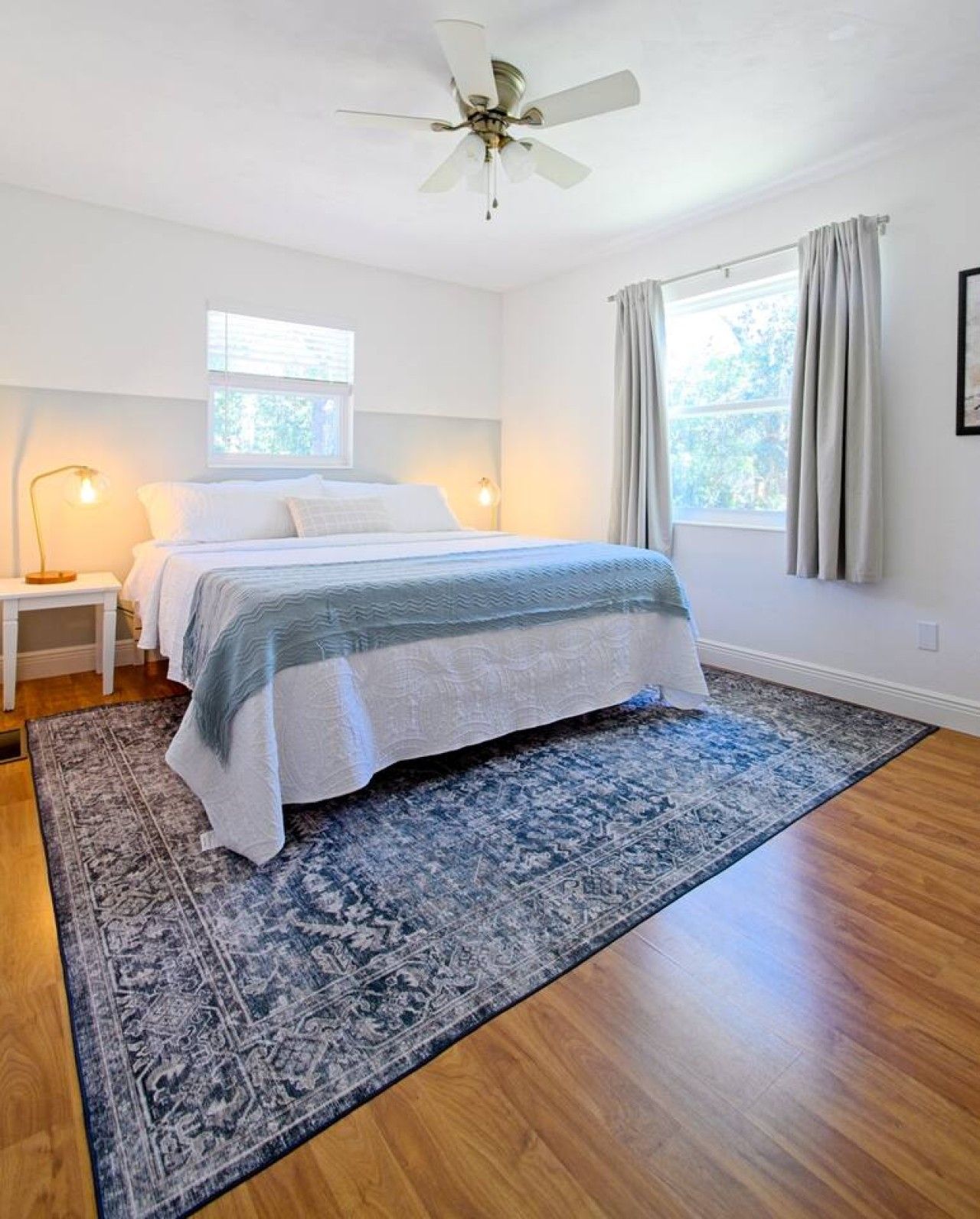A bedroom with a bed, a nightstand, a floor lamp, a ceiling fan, a window with curtains, and a patterned rug.