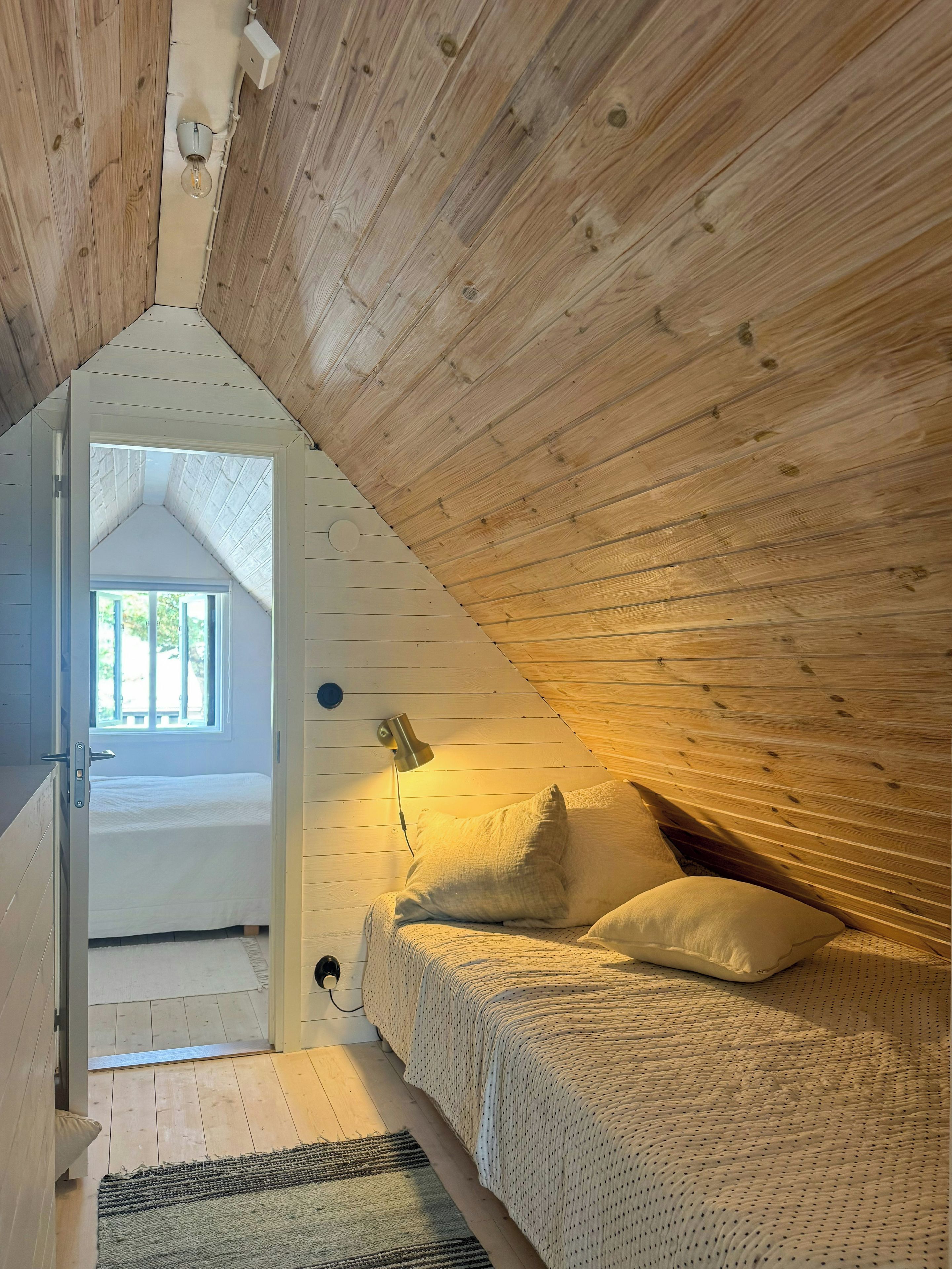 Un dormitorio con techo inclinado, cama con almohadas, lámpara de noche y ventana.
