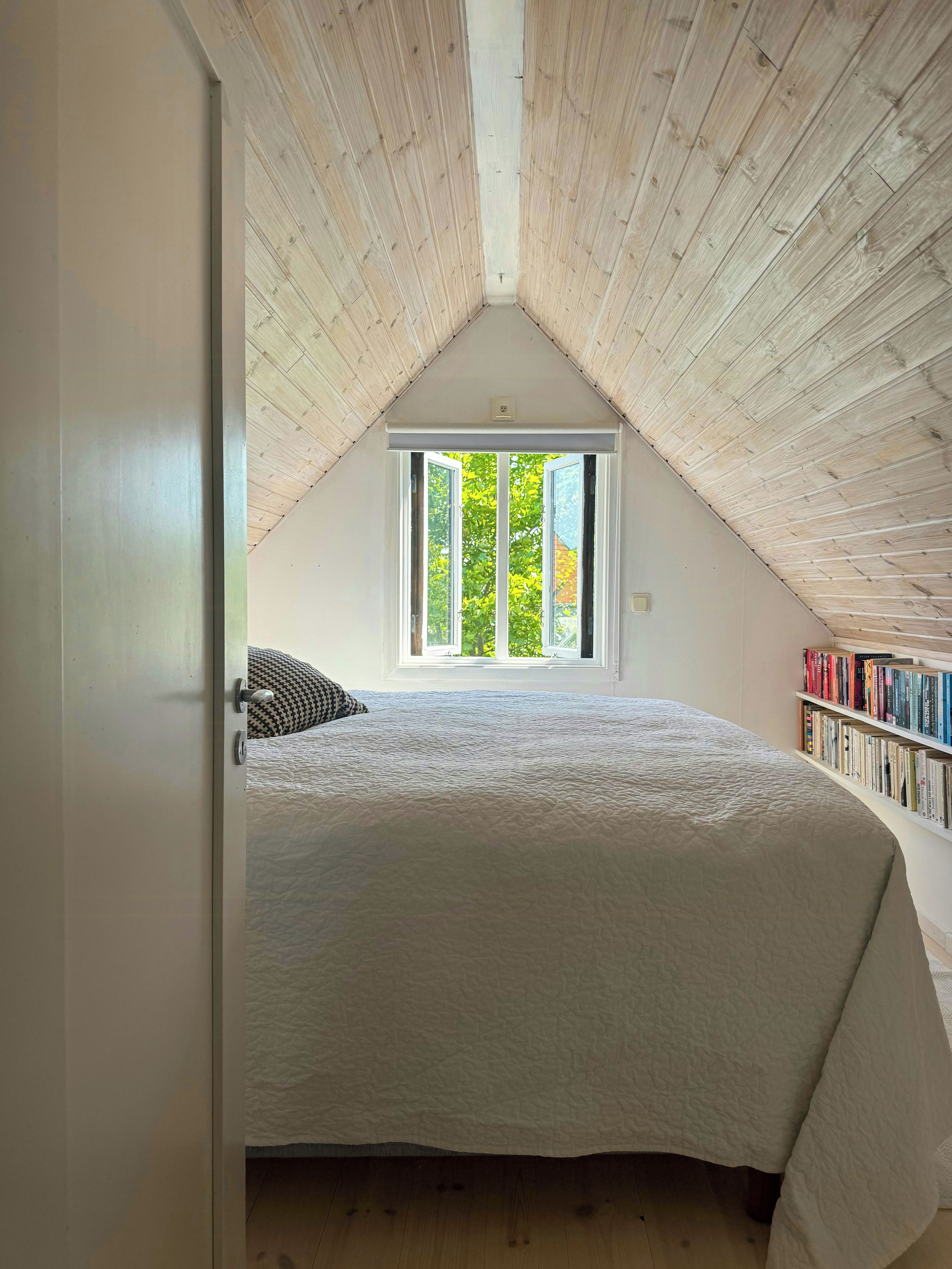 Une chambre avec un plafond en pente, une grande fenêtre, un lit et une bibliothèque.