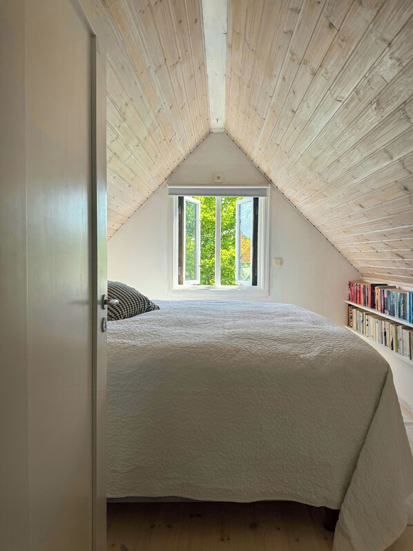 A bedroom with a sloped ceiling, a large window, a bed, and a bookshelf.
