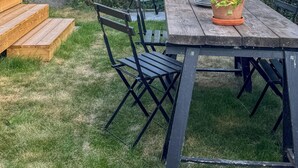 Taula de fusta amb caires, una planta en test i una barbacoa al pati d'una casa, amb tanca de fusta i vegetació.