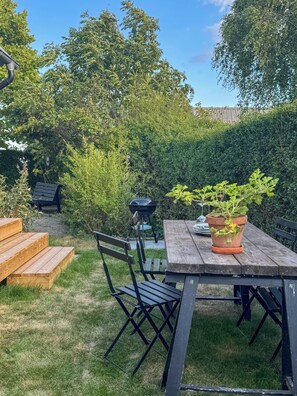 A wooden table with chairs, a potted plant, and a barbecue grill in a backyard with a wooden fence and greenery. - Renovated house with garden in Österlen (Skåne län)