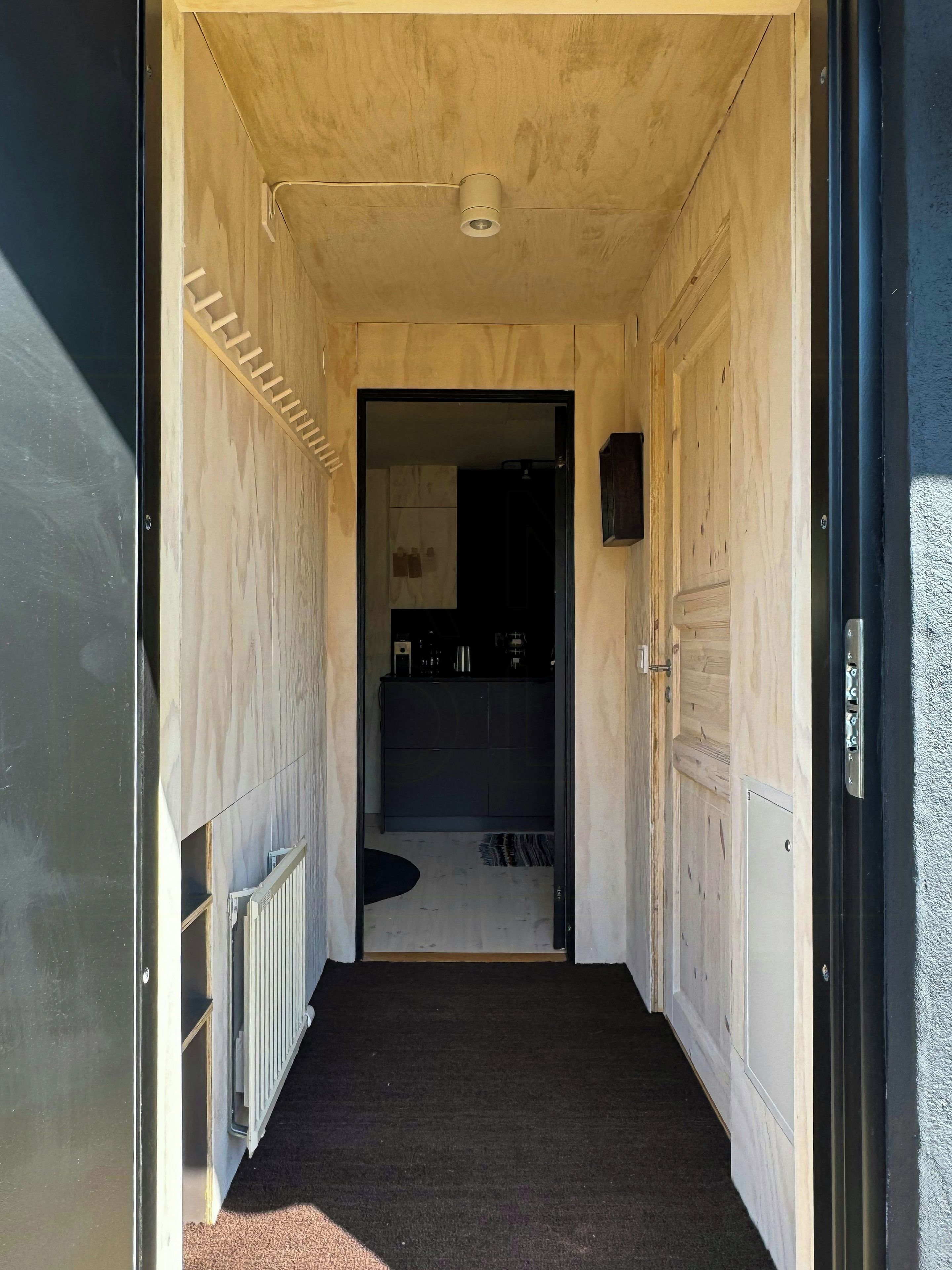 Un couloir étroit, avec un radiateur sur le côté gauche, une moquette sombre et une porte donnant sur une pièce à l’intérieur sombre.