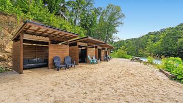 A beachside wooden structure with seating and a picnic table, surrounded by greenery and a clear sky.