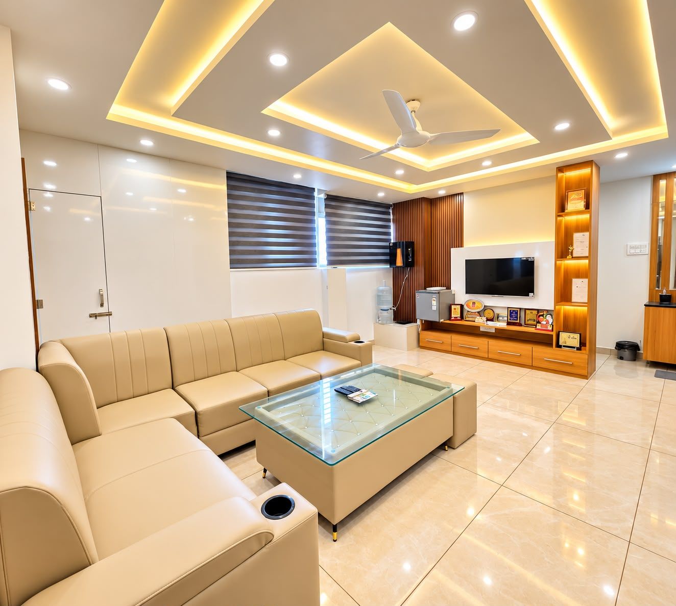 A modern living room with a beige sofa, a glass coffee table, recessed ceiling lighting, a flat-screen TV, and a wooden entertainment unit with awards.