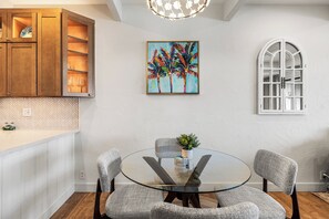 Un comedor con una mesa de vidrio rodeada de cuatro sillas, un cuadro con palmeras y un ventanal con marco blanco.