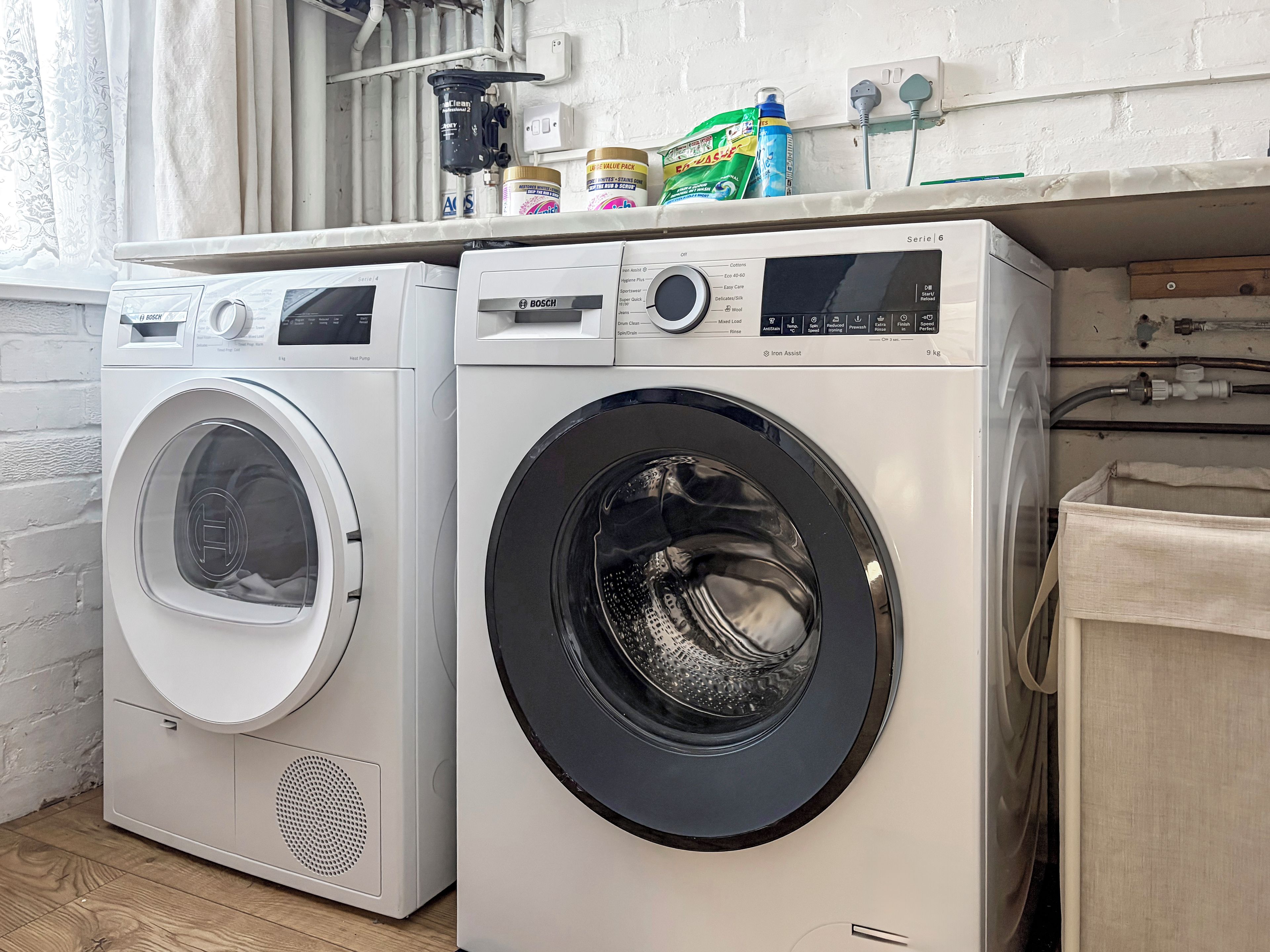 Two front-loading washing machines in a laundry area with a white brick wall and a window with curtains.