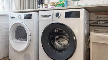 Two front-loading washing machines in a laundry area with a white brick wall and a window with curtains.