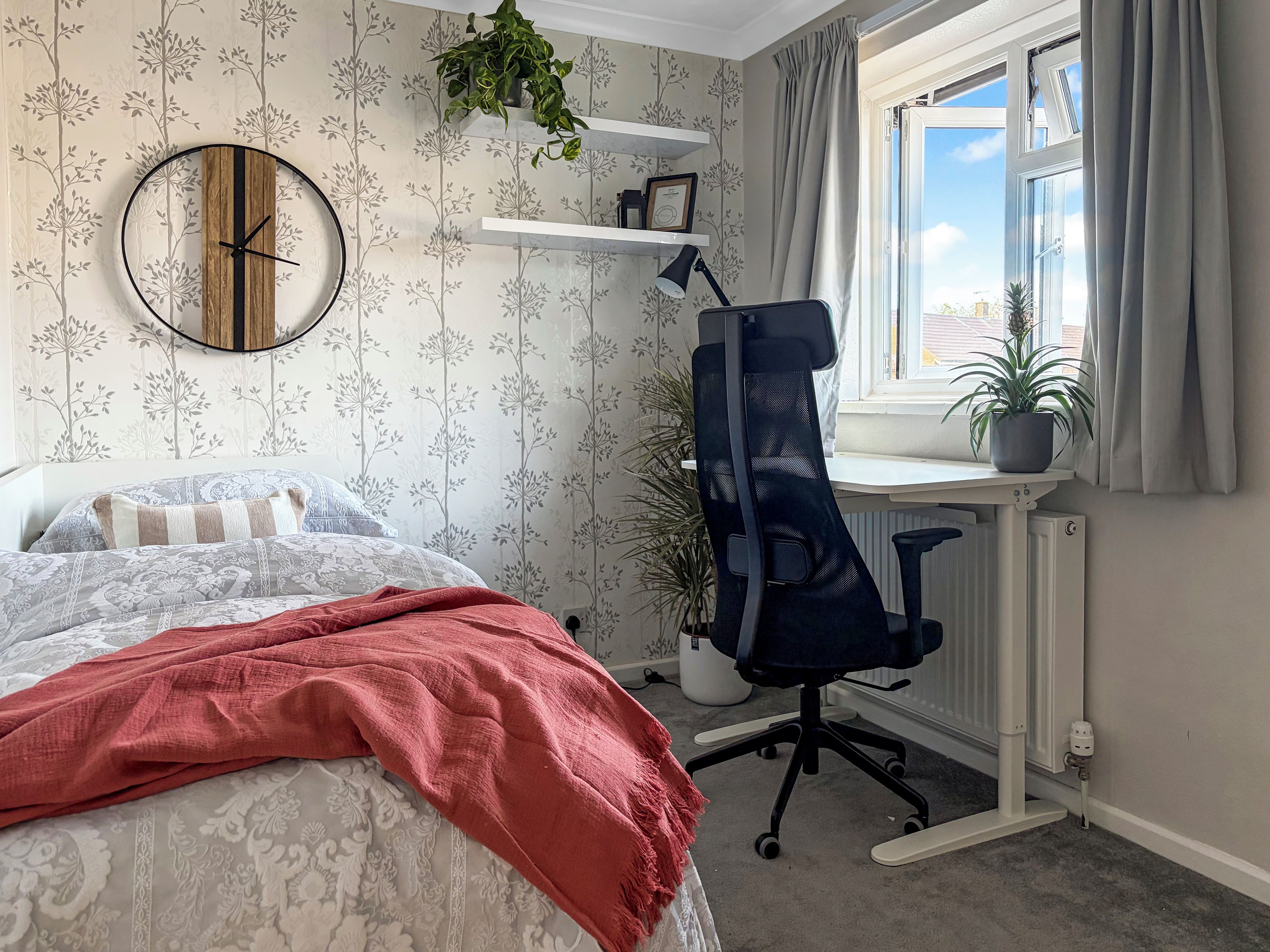 A bedroom with a bed, a desk, a chair, a window with curtains, and a wall clock.