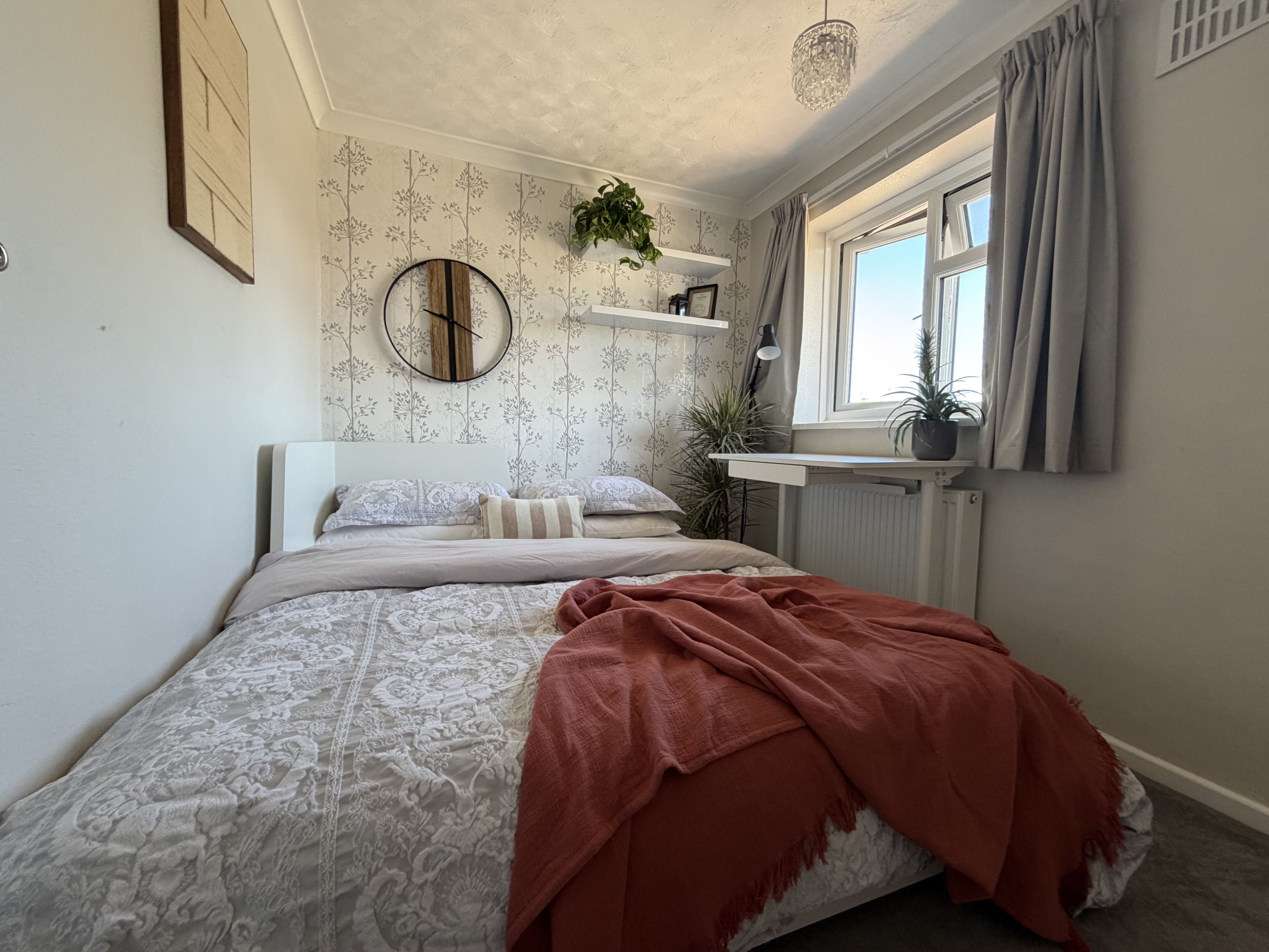 Un dormitorio con cama, un reloj de pared redondo, una ventana con cortinas, un radiador y una planta en maceta.
