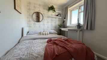 Un dormitorio con cama, un reloj de pared redondo, una ventana con cortinas, un radiador y una planta en maceta.