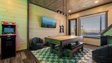 Una habitación con techo de madera, un televisor montado, una mesa, sillones de mimbre y una ventana con persianas.