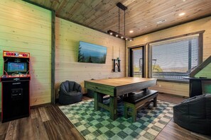 Una habitación con techo de madera, un televisor montado, una mesa, sillones de mimbre y una ventana con persianas.