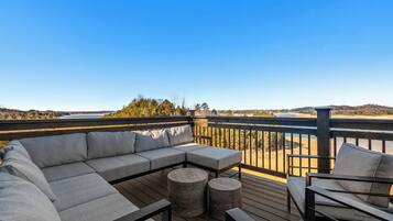 An outdoor deck with a sectional sofa, two round stools, and a chair, overlooking a body of water and distant landscape.