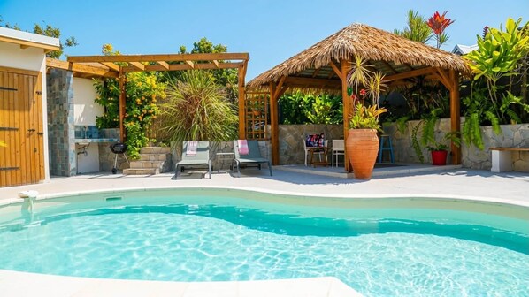 Una piscina con un porche de madera y una estructura de techo de paja, rodeada de plantas en macetas y mobiliario exterior.