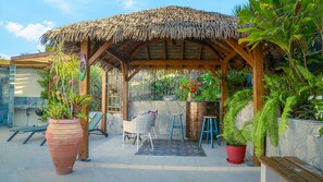 Un espacio de estar al aire libre cubierto, con techo de paja, plantas en macetas y una pared de piedra.