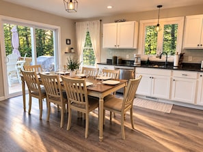 Un comedor con mesa y sillas de madera, una cocina con armarios blancos y vistas a zonas verdes.