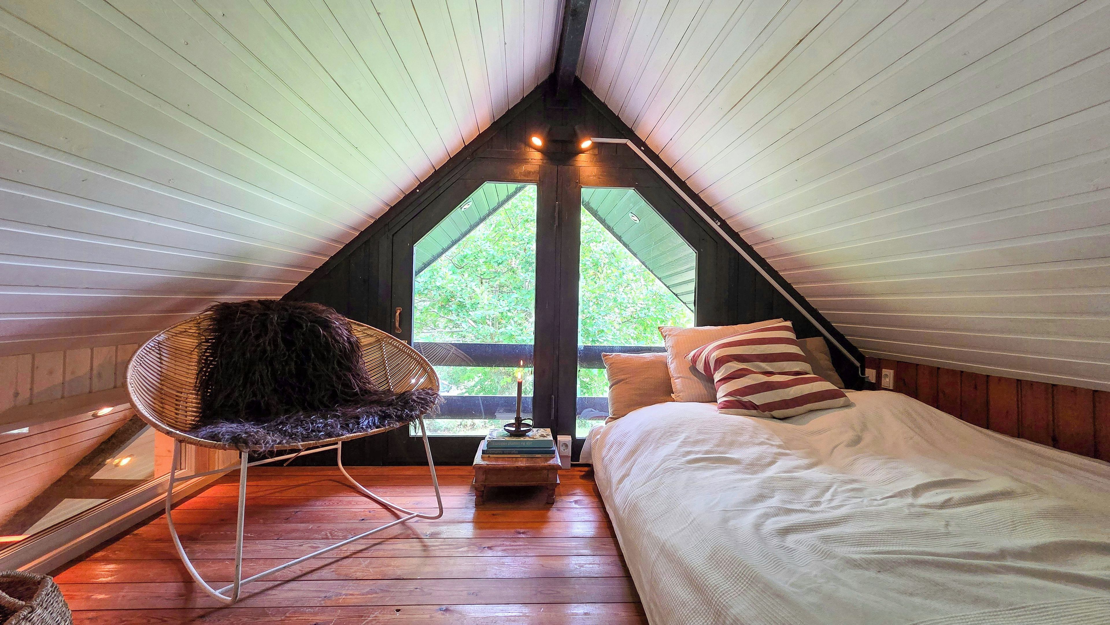 A cozy attic bedroom with a bed, a chair, and a view of greenery outside.
