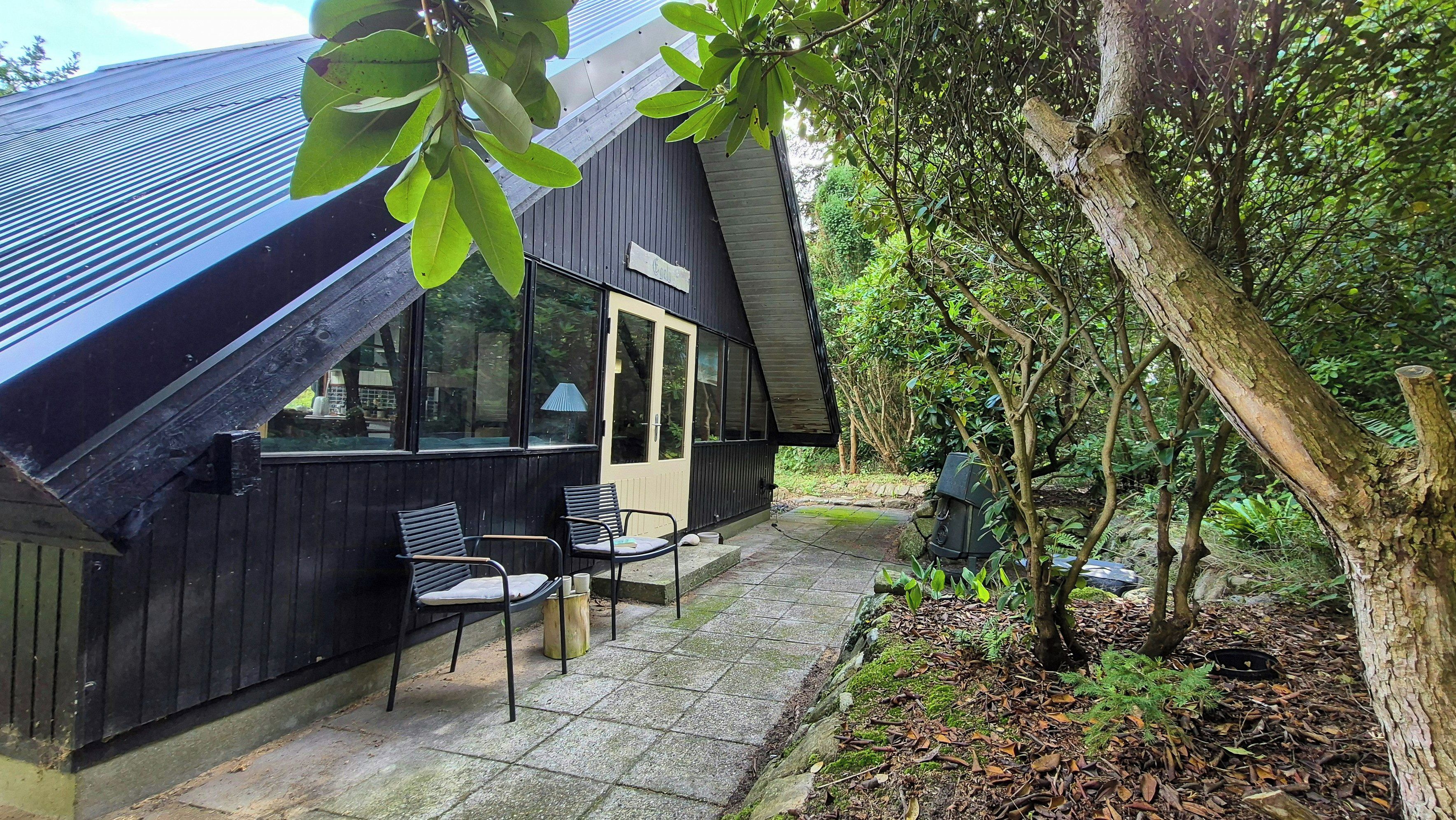 Een zwart gebouw met een schuin dak, een klein terras met twee stoelen, en een bord waarop staat 'Cottage'.