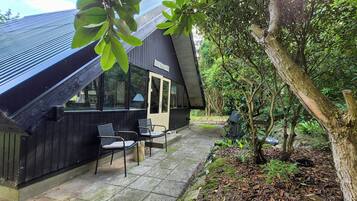 Een zwart gebouw met een schuin dak, een klein terras met twee stoelen, en een bord waarop staat 'Cottage'.