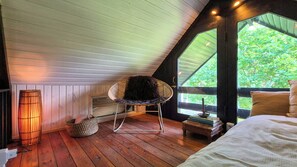 Een gezellige kamer met een schuine kap, een rieten stoel, een nachtkastje met boeken en uitzicht op groen.