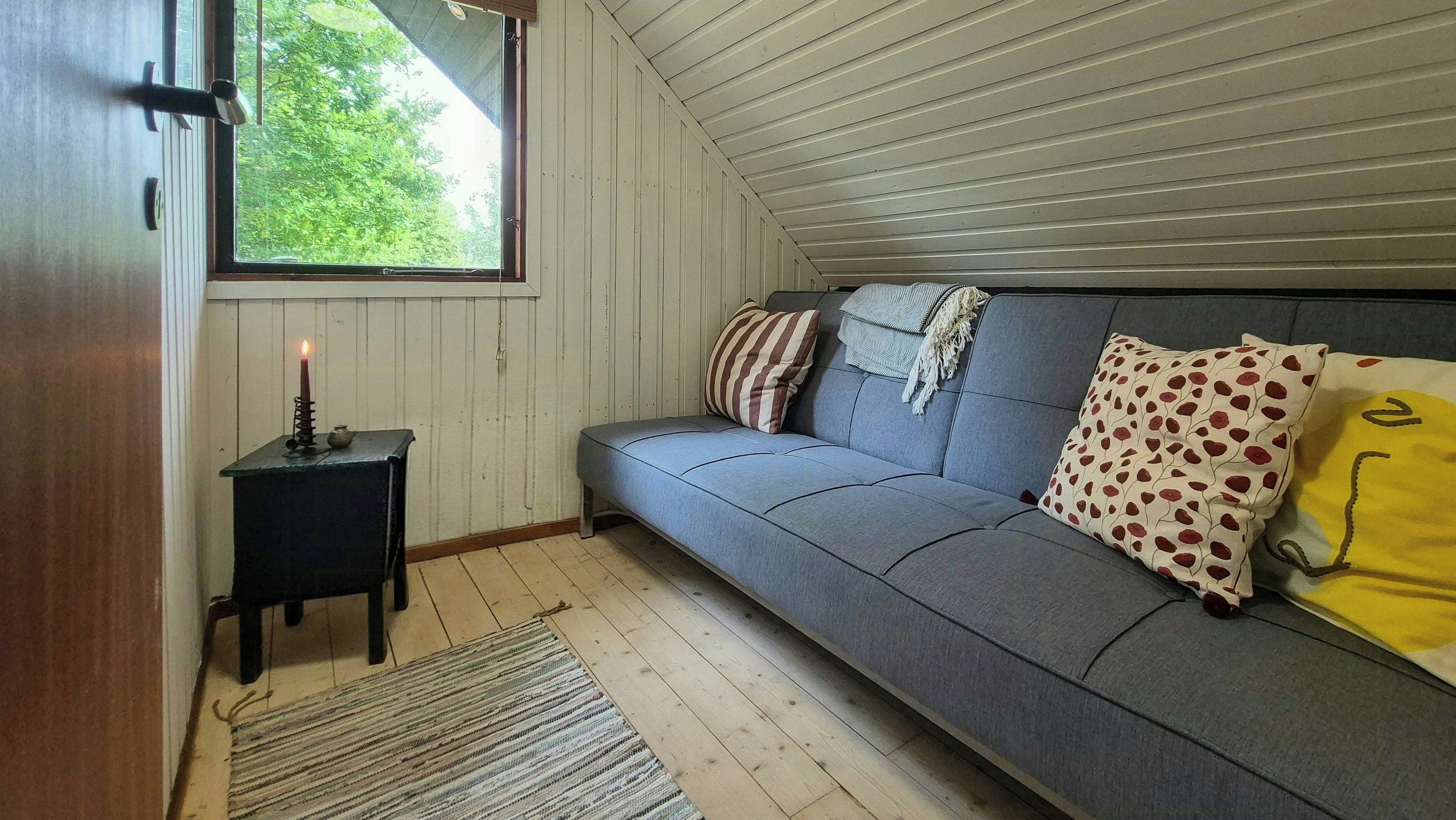 Ein gemütliches Zimmer mit einem grauen Sofa, einem kleinen Tisch mit Kerze und Blick auf Grün durch ein Fenster.