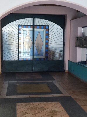 Uma porta dupla com um desenho geométrico em vitral, dando acesso a uma área de entrada revestida com azulejos.