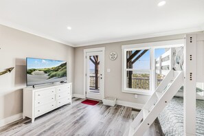 Ein modernes Interieur mit einem Flachbildfernseher, einer weißen Kommode, einer roten Fußmatte und Blick auf den Strand durch ein großes Fenster.