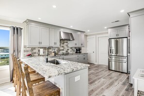 A modern kitchen with white cabinets, a stainless steel refrigerator, and a marble countertop. - Gorgeous Beachfront Luxury 6 BR Home | Elevator | Pet Friendly | Free Activities Including Free Golf (Oak Island)