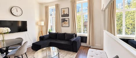 A modern living room with a black sectional sofa, a glass coffee table, a dining area with a table and chairs, and large windows with curtains.