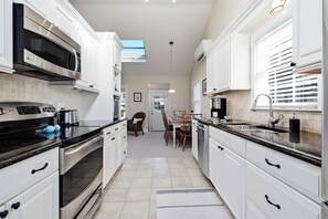 A modern kitchen with white cabinetry, stainless steel appliances, and a dining area with a table and chairs. - Moody Surfer - Coastal Retreat (Wells)