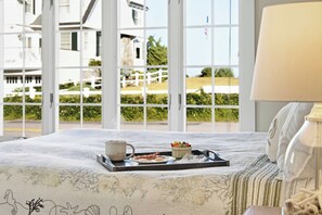 A bed with a tray of breakfast items, a lamp, and a view of a white house through large windows. - Moody Surfer - Coastal Retreat (Wells)