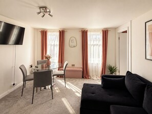 A modern living room with a black sofa, a glass table with chairs, a flat-screen TV, and a large window with curtains. - Stylish Fitzrovia 1BR Near Oxford Street (Greater London)