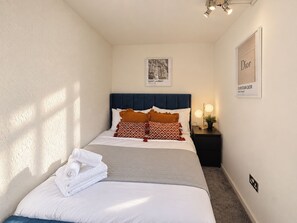 A neatly made bed with white linens and a folded towel, a dark blue headboard, a bedside table with a lamp, and a framed picture on the wall. - Stylish Fitzrovia 1BR Near Oxford Street (Greater London)