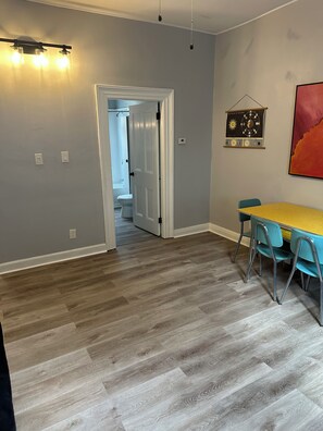 Ein Zimmer mit Holzboden, einem Esstisch mit Stühlen, einer zur Badezimmertür führenden Öffnung und einer an der Wand befestigten Leuchte.