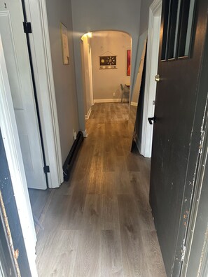 A hallway with a wooden floor, a door on the right, and a wall with a clock and a framed picture.