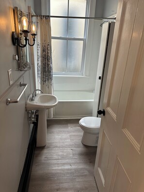 Ein Badezimmer mit weißem Waschbecken, WC und Dusche. Ein Fenster mit Milchglas und ein Vorhang sind vorhanden.