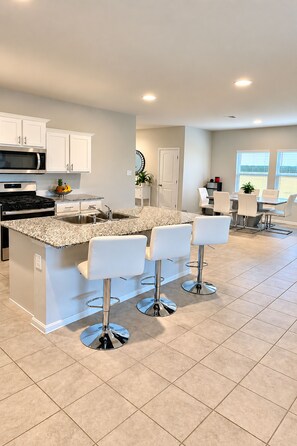 A modern kitchen with a central island, bar stools, and a dining area with a table and chairs. - Upscale 3BR - 2.5 Bathroom Home with 2 Living Rooms + Gym & Pool (Cresson)