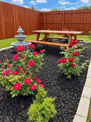 Un jardin avec une clôture en bois, une fontaine en pierre, une table de pique-nique et des arbustes à fleurs rouges.
