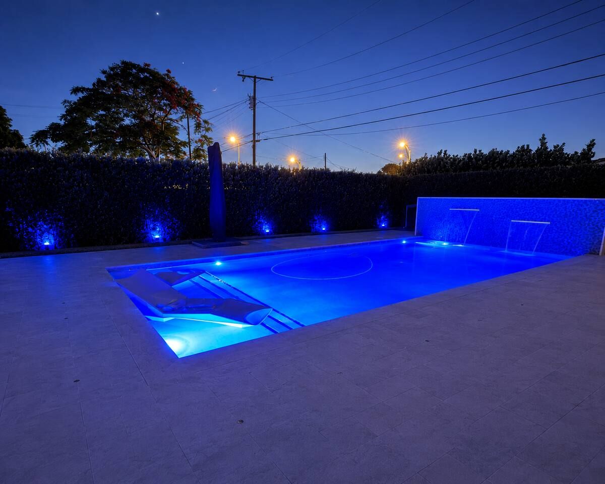 Une piscine dont le bord est illuminé de lumière bleue, entourée d’une haie et bordée de poteaux électriques.