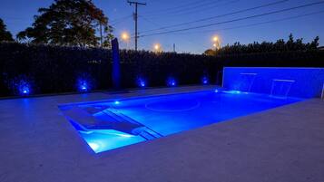 Une piscine dont le bord est illuminé de lumière bleue, entourée d’une haie et bordée de poteaux électriques.