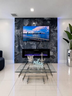 A modern living room with a TV mounted on a marble wall, a fireplace below, and a glass coffee table with a neon sign.