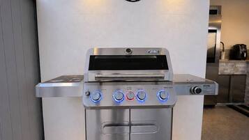 Un barbecue d’extérieur en acier inoxydable, doté d’un écran numérique et de plusieurs boutons de commande, placé devant une enseigne fixée au mur sur laquelle on peut lire « ZONE BARBECUE ».