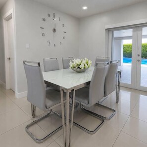 A dining area with a white table, grey chairs, a clock on the wall, and a view of a swimming pool through a glass door.
