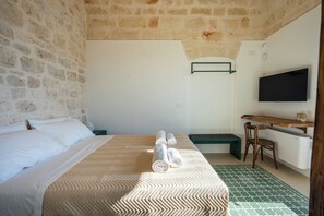 A hotel room with a stone wall, a bed with white linens, a bench, a desk, and a television. - Dimora Bellarosa - A Timeless Masseria with Infinity Pool (Puglia)