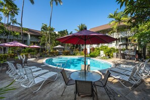 A pool area with lounge chairs, umbrellas, and a glass table. - Kona Islander Inn#251: Downtown Kona, Remodeled! (Kailua-Kona)
