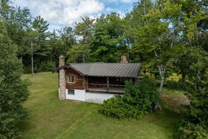 Exterior view of a rustic log cabin with a covered porch set amidst a grassy wooded clearing. - Escape to a Cozy Cabin in the Weston Woods (Weston)