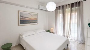 Et hotelværelse med en seng, et natbord med lampe, en vægmonteret aircondition, et vindue med gardiner og et billede på væggen.